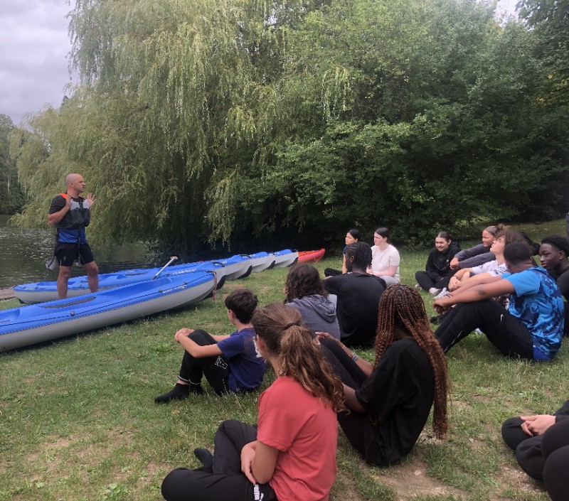 Groupe d'élève assis face au lac, écoutant les instructions du professeurs pour manipuler les canoës
