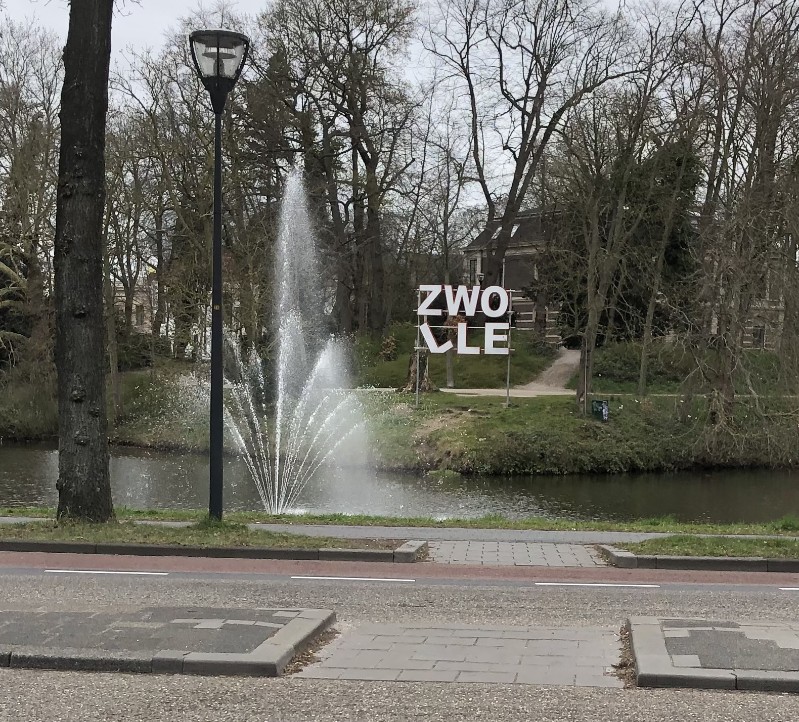 L'image montre un panneau sur lequel "Zwolle" est écrit en deux parties « Zwo » et « lle », près d'un canal de la ville et en arrière-plan d'un jet d'eau. L'arrière-plan présente une scène avec des arbres. 