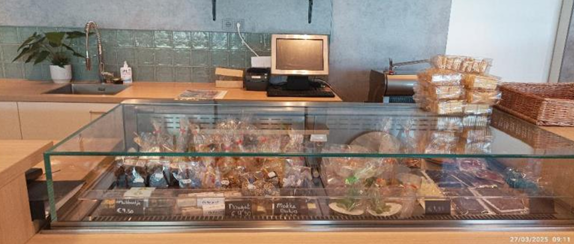 Cette photo montre l'espace dédié à la vente de produits de boulangerie et de pâtisserie au Deltion College. Les produits sont disposés dans une vitrine réfrigérée et étiquetés. L'espace est bien organisé, avec un comptoir et une caisse enregistreuse en arrière-plan.