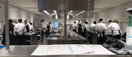 Sur cette photo, des élèves en uniforme travaillent dans une cuisine professionnelle d'application, avec des équipements de cuisine de haut niveau. Plusieurs étudiants sont impliqués dans la préparation des plats sous la supervision d'enseignants. 