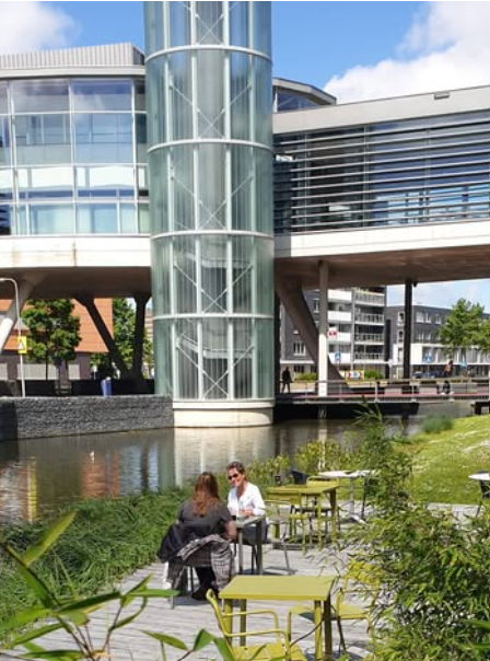 L'image présente l'extérieur moderne et spacieux du Deltion College, avec des fenêtres et des structures transparentes et un canal qui entoure l'établissement. 