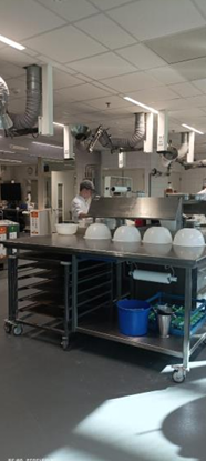 Photo d'une cuisine d'application moderne, équipée de tables de travail en métal, d'étagères à roulettes et de bacs blancs alignés. Des systèmes de ventilation sont visibles au plafond, et un chef est en train de travailler.