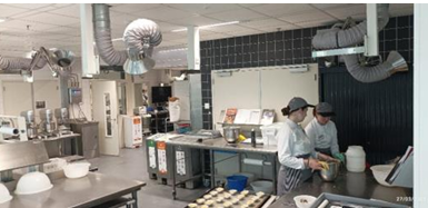 L'image montre une cuisine moderne d'application où les étudiants pratiquent la préparation des plats du déjeuner. L'espace est équipé de plusieurs stations de travail et d'appareils de cuisson professionnels.